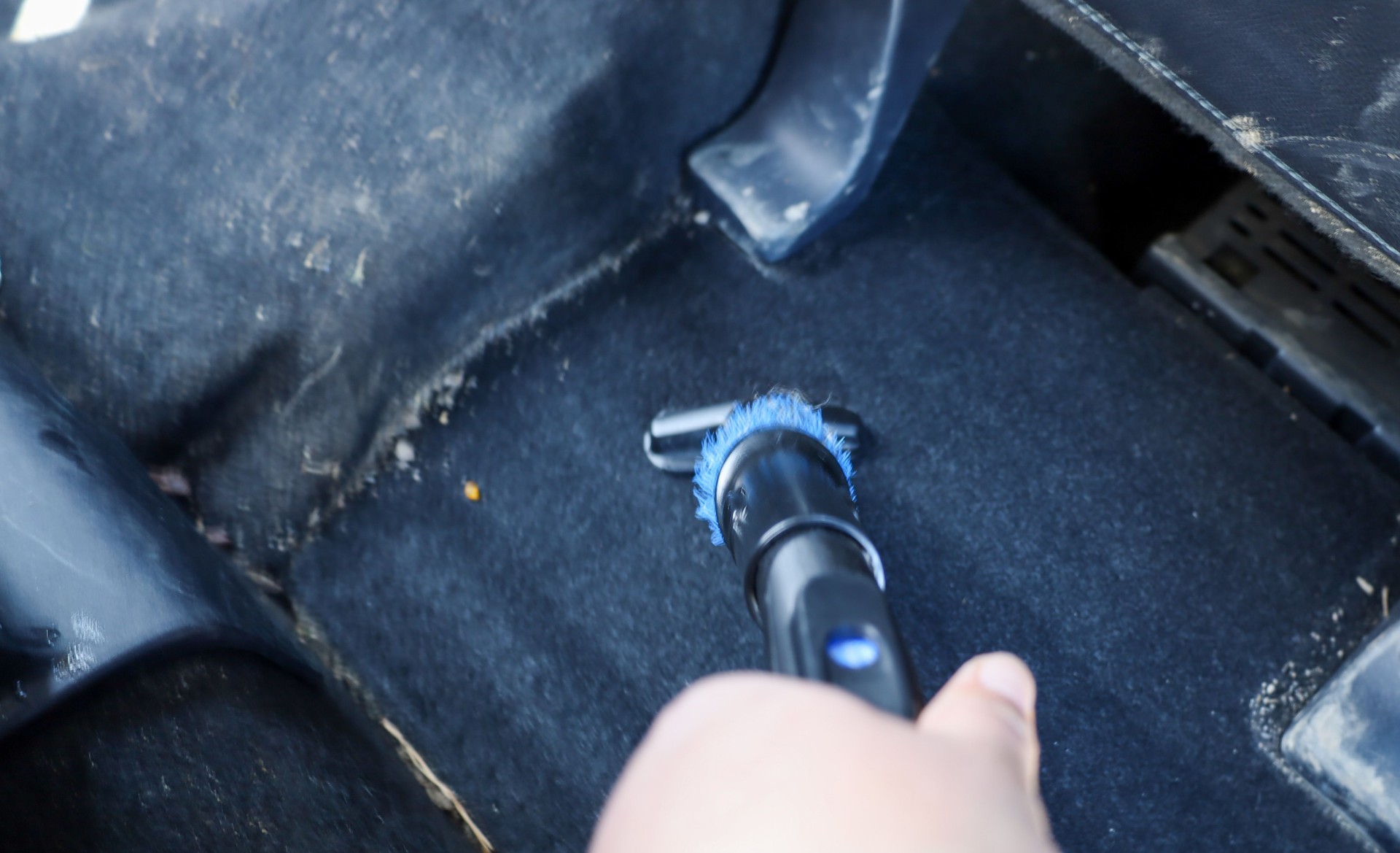 A close-up photograph shows a hand vacuuming a dirty car carpet. A handheld vacuum cleaner with a blue brush removes dirt and dust from the black carpet.